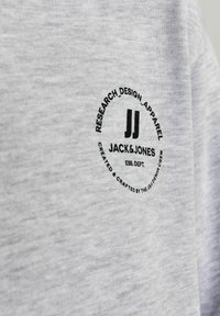 Szürke pamut póló, kerek fekete logóval, amelyen a "JACK & JONES" felirat, valamint a "RESEARCH_DESIGN_APPAREL" és "ESS. DEPT." kiegészítések láthatók.