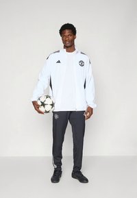 Bílá sportovní bunda s černými akcenty, černým logem, doplněná šedými kalhotami s bílými pruhy, držící černo-bílý fotbalový míč.