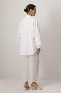 Hvid lagdelt bluse med gennemsigtig stof og klokkeærmer, parret med en lang, let gennemsigtig hvid nederdel. Spidse beige hæle fuldender outfittet.