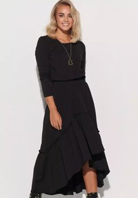 Blondynka uśmiechająca się, ubrana w czarną bluzkę z długim rękawem, asymetryczną czarną spódnicę, czarne botki i złotą naszyjnik z wisiorkiem, stojąca przed białą ścianą.