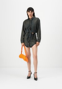 Model într-o cămașă din denim legată în nod și fustă mini, ținând o geantă portocalie strălucitoare, purtând pantofi cu toc înalt negri, pe un fundal alb.