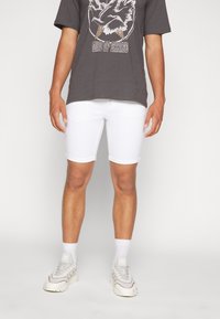 Pantalones cortos de mezclilla blanca con dobladillos doblados, combinados con una camiseta gris con un estampado gráfico. El modelo lleva zapatillas ligeras con calcetines blancos.