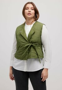 Grüner gesteppter Weste mit einer Bindeschließe, mit Kragen und Druckknopf. Getragen über einem weißen Hemd, mit dunklen Jeans. Glatte Stofftextur.