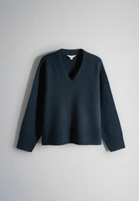 Tmavě námořnicky modrý pletený svetr s dlouhými rukávy a výstřihem do V visící na světle šedém pozadí.