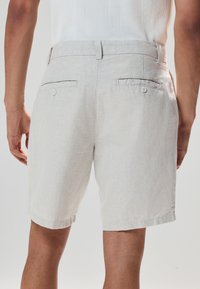 Shorts en lin gris clair avec une coupe décontractée, ourlet roulé et deux poches arrière à boutons. Texture lisse et design minimaliste.