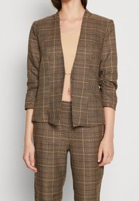 Blazer marron à carreaux avec un design sans col, des poches avant et des manches ajustées, assorti à un pantalon à motifs assortis.