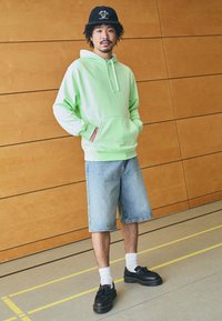 Hoodie verde claro com bolso frontal, calções de ganga, meias brancas e sapatos pretos com solas grossas. Fundo minimalista com painéis de madeira.