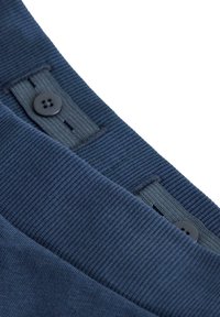 Tessuto blu navy con una texture a coste, caratterizzato da dettagli con bottoni e accenti a tracolla. Una prospettiva ravvicinata del bordo del capo mette in risalto le cuciture e la forma.