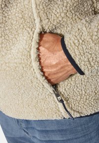 Cream-colored fleece jacket with a textured, fluffy surface. Zippered pockets and a navy accent at the cuff. Hand placed in pocket.