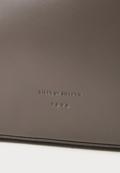 Donkerbruine leren tas met het verdiept aangebrachte merk K tekst "TIGER OF SWEDEN" en "1903" op een glad oppervlak. Eenvoudig, rechtlijnig ontwerp.