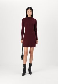 Vestido ribatejado borgonha com mangas longas e silhueta ajustada, apresentando um acabamento em ponta. Modelo segura uma bolsa preta, vestindo botas pretas.