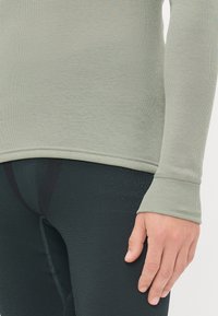 Grønn langermet undertrøye med ribbet tekstur, med rund nederkant og tommelhull. Svart tettsittende leggings med subtile mønsterdetaljer.