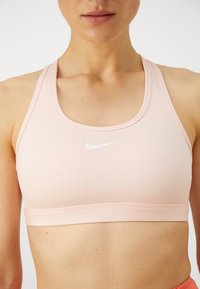 Sujetador deportivo rosa claro, fabricado con material sintético suave, que presenta un escote redondeado y un pequeño logo blanco de Nike centrado en la parte frontal.