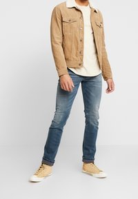 Man som bär en ljusbrun kordjacka med vit fleecekrage, en krämfärgad t-shirt, blå uppvikta jeans och gula högstövlar, stående mot en vit vägg.