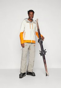 Ski jacket in white and orange, with a zip front and hood. Beige ski pants with pockets. Black ski boots. Ski in hand, standing pose.