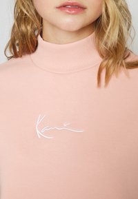Femme portant un pull à col roulé côtelé rose clair avec le logo "Kani" en cursive blanche brodé sur la poitrine.