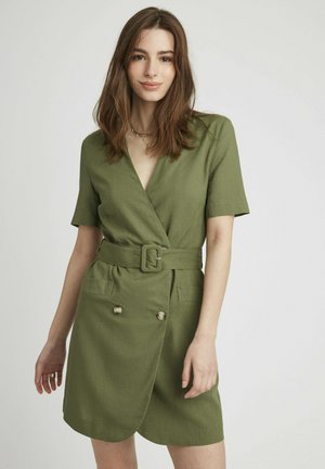 Robe portefeuille vert olive avec des manches courtes, taille ceinturée, deux poches avant et accents de boutons. Fabriquée en tissu texturé.