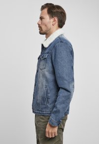 Giubbotto di jeans blu medio con colletto in pelliccia sintetica bianca, dotato di tasche laterali e chiusura a bottoni, mostrato di lato.