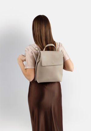 Donna con lunghi capelli castani, indossa una camicia beige e una gonna marrone scuro, mentre sistema gli spallacci in pelle grigio chiaro dello zaino sulla schiena.
