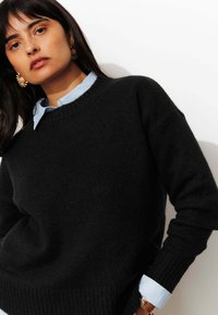 Pull en maille noire avec un col rond et un ourlet côtelé. Porté par-dessus une chemise à col bleu clair. Comptant de longues manches et une texture douce.