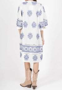 Robe blanche avec broderies florales bleues, manches trois-quarts, coupe ample et finition en dentelle à l'ourlet. Portée avec des bottes de cowboy beiges.