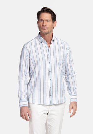 Man die een lichtblauw-wit gestreept overhemd met knopen en een witte broek draagt, staand tegen een effen achtergrond.