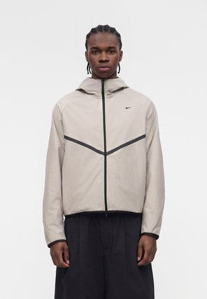 Ung mand med flettet hår iført en beige Nike-jakke med sort lynlås og chevrondetalje, kombineret med løse sorte bukser mod en ensfarvet baggrund.