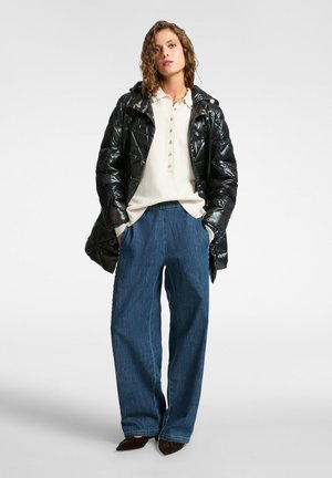 Giacca puffer nera con finitura lucida, chiusura con bottoni, indossata sopra un maglione beige e pantaloni di denim blu larghi. Scarpe marroni scure.