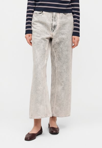 Jean gris clair à jambes larges avec une finition délavée, doté de poches avant et de détails de couture visibles. Associé à des chaussures plates foncées.