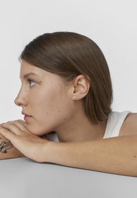 Perfil de una persona con cabello castaño medio, vistiendo una blusa sin mangas blanca y un pequeño pendiente de perla. La piel muestra pecas y una textura suave.