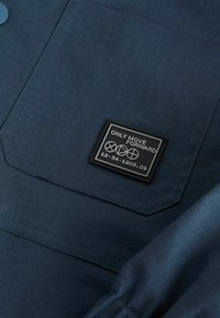 Sötétkék, textúrált felületű anyag, fekete téglalap alakú címkével, amelyen fehér szöveggel ez áll: "CSAK ELŐRE MOZOGJ".