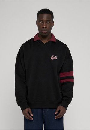 Jeune homme portant un sweat noir avec un col bordeaux, des rayures bordeaux sur la manche droite et un logo "Ecko Unltd" sur la poitrine.