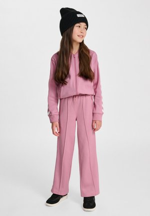 Meisje draagt een roze trainingspak met een jas met ritssluiting en wijde broekspijpen, een zwarte muts en zwarte sneakers, staat en kijkt opzij.