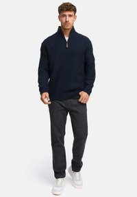 Mand iført en marineblå quarter-zip sweater, mørke jeans og hvide sneakers, der står op imod en ensfarvet lys baggrund.