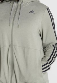 Sweat à capuche zippé vert clair avec des accents gris, doté d'une capuche, de deux poches avant et de trois bandes noires sur chaque manche. Logo Adidas affiché.