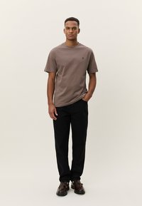 Καφέ βαμβακερό T-shirt με κοντά μανίκια και στρογγυλή λαιμόκοψη, με μικρό λογότυπο. Συνδυάζεται με μαύρο παντελόνι και καφέ παπούτσια.