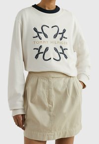 Sudadera blanca con estampado de cuerda en azul marino y texto en dorado "Tommy Hilfiger", combinada con una falda beige con pliegues y cinturilla elástica.
