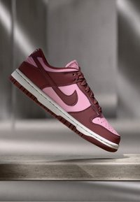 A Nike Dunk sneaker piros és rózsaszín bőrből készült felsőrésszel rendelkezik, fehér középtalppal és rózsaszín talppal. A dizájn jellemzője a perforált lábujj doboz és a swoosh.