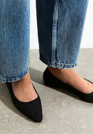 Piedi con ballerine nere a punta abbinate a jeans blu con orlo sfrangiato su un pavimento grigio chiaro.