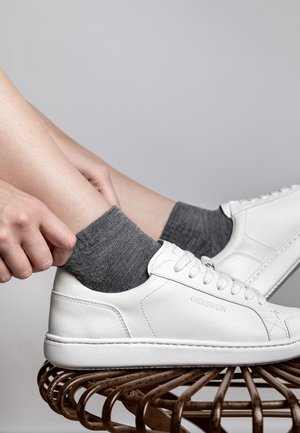 Witte leren sneaker met grijze textuursokken, met een ronde neus, platte zool en vetersluiting. Schoen geplaatst op een geweven bruine kruk.