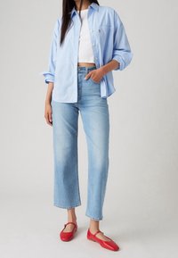 Světlomodrá košile na knoflíky vrstvená přes bílý crop top, v kombinaci se světlomodrými džíny s vysokým pasem a červenými plochými botami se strapcem.