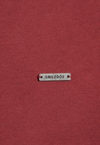 Červená tkanina s žebrovanou texturou, na dolním okraji připevněná kovová visačka s nápisem "SMILODOX".