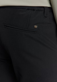 Zwarte broek van een gestructureerde stof, met een achterzak en een hexagonaal zwart-gouden accent. Elastische tailleband voor comfort.