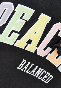 Czarna bluza z kapturem z dużym, wielokolorowym napisem „PEACE” w pastelowych odcieniach. Pod spodem mniejszy, biały napis „BALANCED.” Tkanina wydaje się miękka.