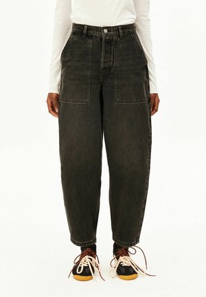 Person trägt schwarze, hochgeschnittene Jeans mit weitem Bein und großen Fronttaschen sowie schwarze Sneakers mit weißen und braunen Schnürsenkeln, steht vor weißem Hintergrund.