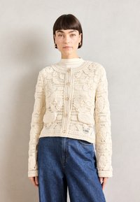 Cremfarbene Häkeljacke mit floralem Muster, runder Ausschnitt, Knopfverschluss und langen Ärmeln, kombiniert mit blauen Jeans.