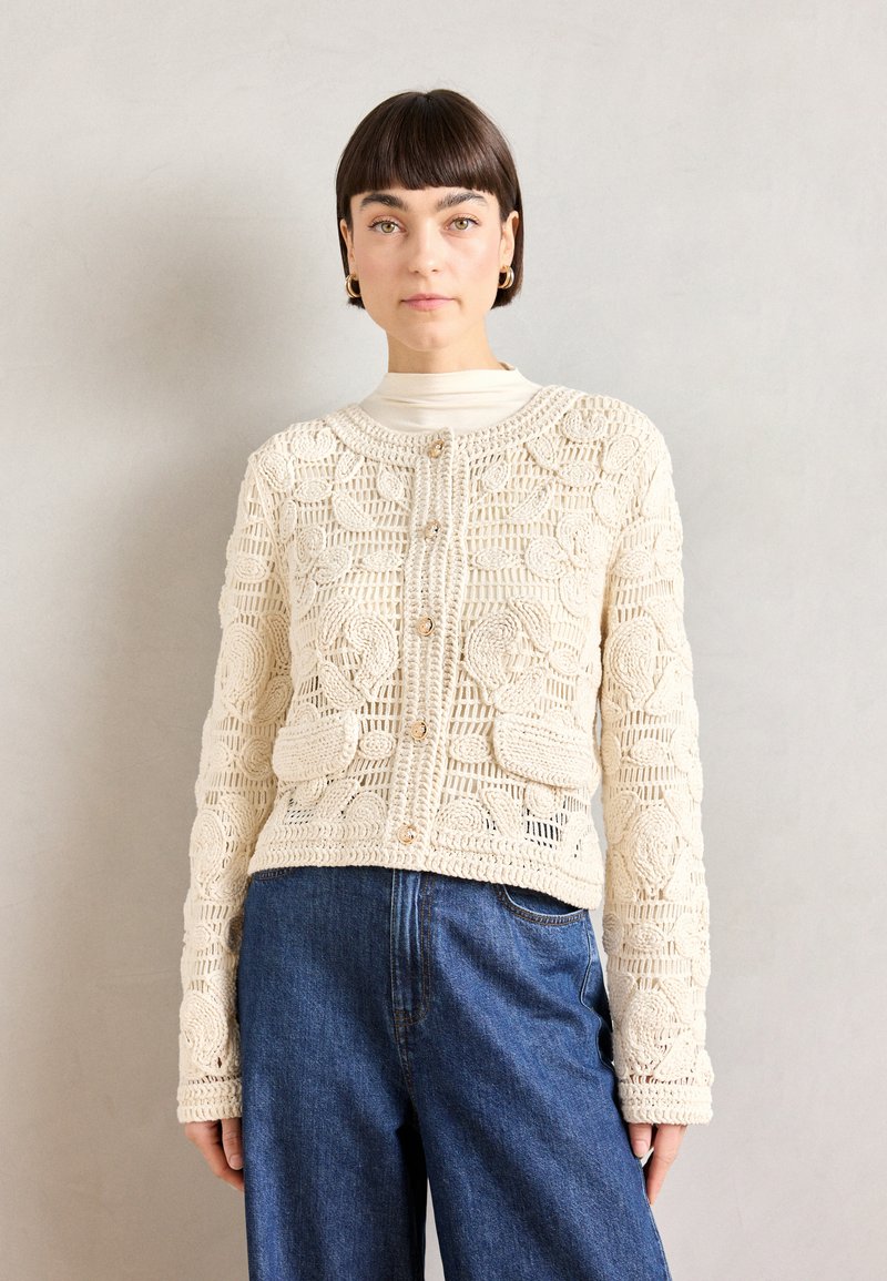 Cremfarbene Häkeljacke mit floralem Muster, runder Ausschnitt, Knopfverschluss und langen Ärmeln, kombiniert mit blauen Jeans.