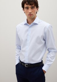Giovane uomo che indossa una camicia da uomo azzurro chiaro, pantaloni blu scuro e una cintura nera, in piedi con le mani in tasca su uno sfondo neutro.