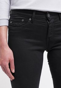 Vaqueros de mezclilla negros con un ajuste slim, que cuentan con bolsillos delanteros, cremallera y herrajes plateados, combinados con un suéter gris claro.