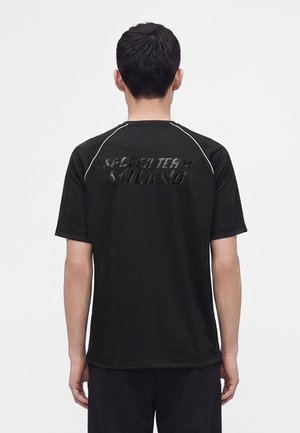 Homme portant une chemise noire à manches courtes avec le texte "Soccer Team Milano" sur le haut du dos, debout devant un fond blanc uni.